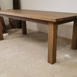 Mariusz isiu - Prostokątny stół z drewna o ciepłym, brązowym odcieniu, z grubym blatem i solidnymi nogami. W tle widoczne deski, co sugeruje warsztat stolarski.