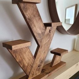 Mariusz isiu - Brązowy, drewniany kwietnik o geometrycznym kształcie na tle ściany z okrągłym lustrem. Widoczne słoje drewna i detale wykonania. Dekoracja wnętrza.