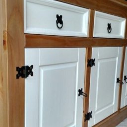 Mariusz isiu - Drewniana komoda z białymi frontami i czarnymi, stylizowanymi uchwytami. Widoczne słoje drewna i detale wykończenia. Ujęcie z boku, podkreślające głębię mebla.