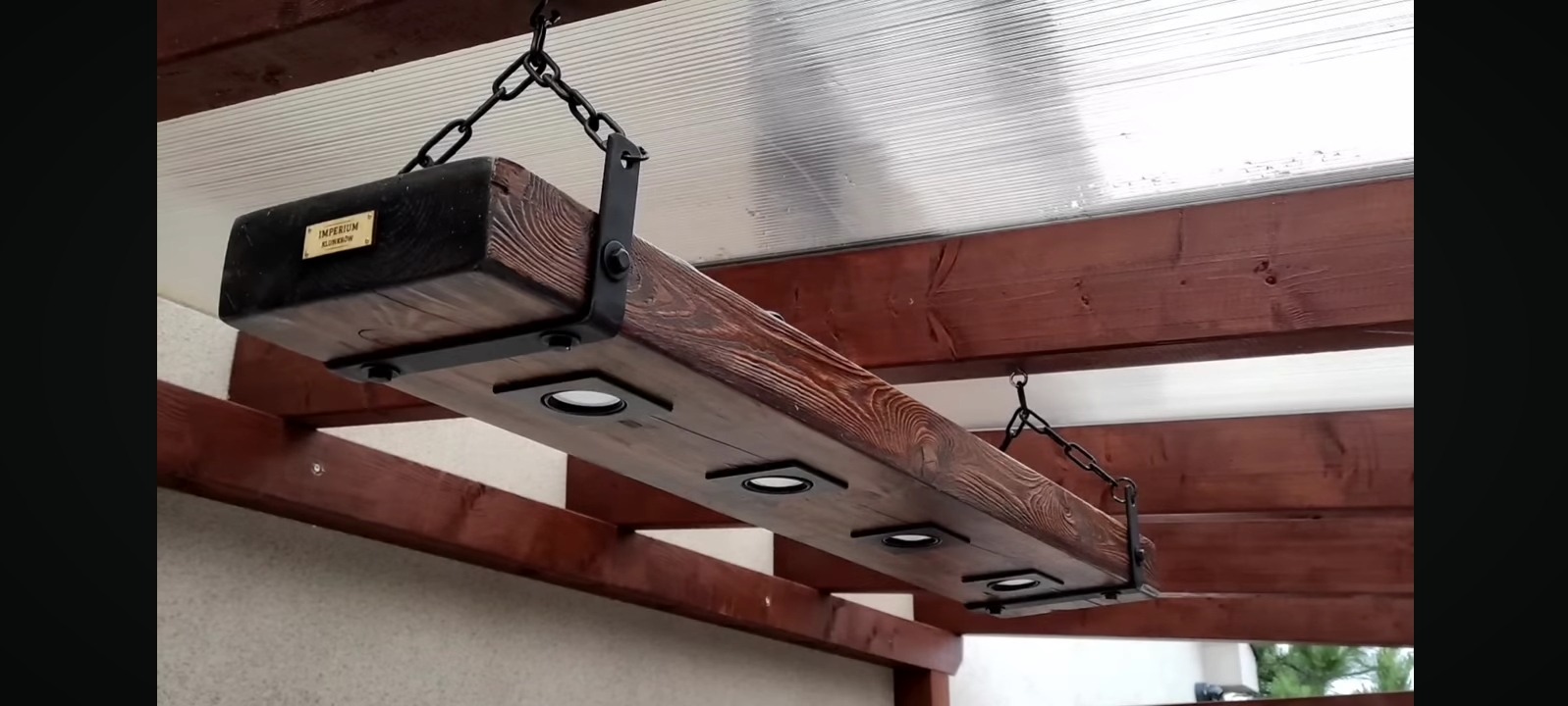 Drewniana belka z oświetleniem LED, zawieszona na łańcuchach pod zadaszeniem. Widoczne detale wykończenia i metalowe elementy mocujące.