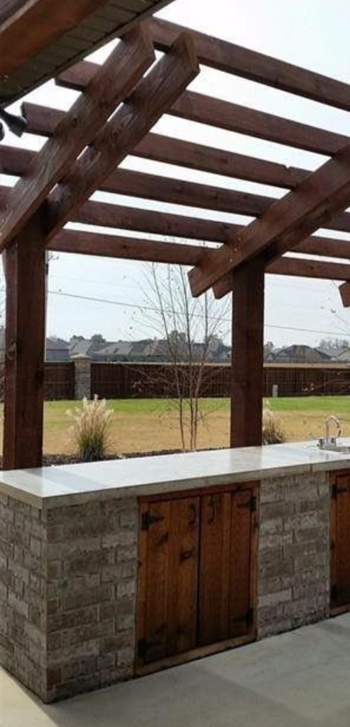 Zadaszona, zewnętrzna kuchnia z blatem i szafkami z drewna i cegły, z widokiem na trawnik i domy w tle. Elementy drewniane w kolorze ciemnego brązu.