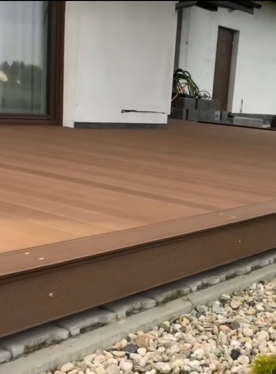 Nowoczesny taras z desek kompozytowych w odcieniach brązu, z widocznymi detalami wykończenia i fragmentem elewacji budynku w tle. Widok z niskiej perspektywy.
