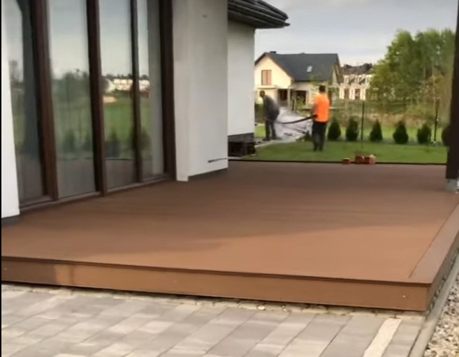 Nowoczesny taras z deski kompozytowej w Żarówce. Widoczne prace wykończeniowe i fragment domu z dużymi oknami. Minimalistyczny design i estetyczne wykończenie.