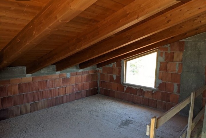 Surowe poddasze z drewnianym stropem i oknem. Ściany z czerwonej cegły, surowy beton na podłodze. Widok na wnętrze w trakcie prac wykończeniowych.