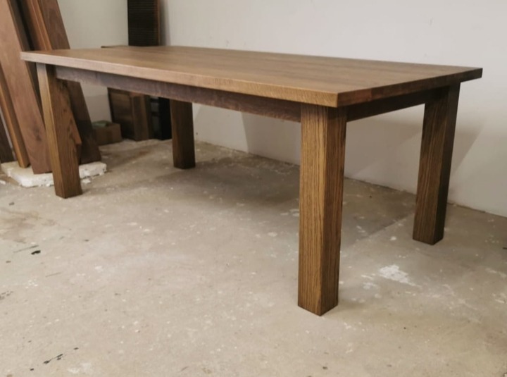 Prostokątny stół z drewna o ciepłym, brązowym odcieniu, z grubym blatem i solidnymi nogami. W tle widoczne deski, co sugeruje warsztat stolarski.