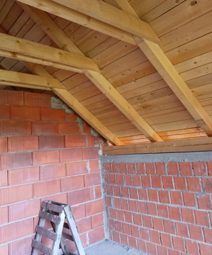 Surowy stan poddasza z drewnianą konstrukcją dachu i murowanymi ścianami z czerwonej cegły. Widoczna drabina oparta o ścianę. Ujęcie z dołu, podkreślające strukturę materiałów.