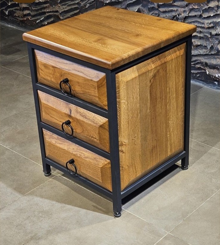 Drewniana szafka nocna z trzema szufladami, w stylu industrialnym, z metalową ramą i okrągłymi uchwytami, na tle ściany z kamienia dekoracyjnego.