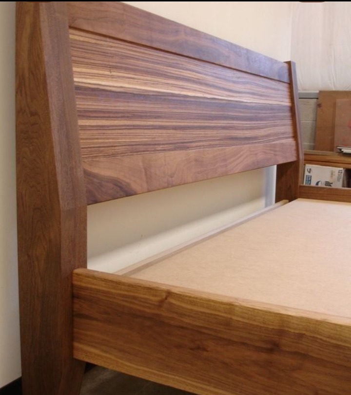 Drewniane łóżko z wezgłowiem o wyraźnym usłojeniu, widoczne detale konstrukcyjne i naturalny odcień drewna. Minimalistyczny design, ciepłe oświetlenie.