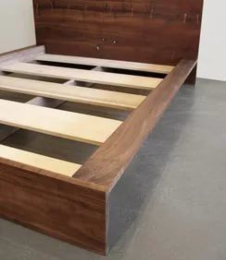 Drewniane łóżko z niskim wezgłowiem i ramą z litego drewna, widoczne stelaże pod materac. Minimalistyczny design, ciepły odcień drewna.