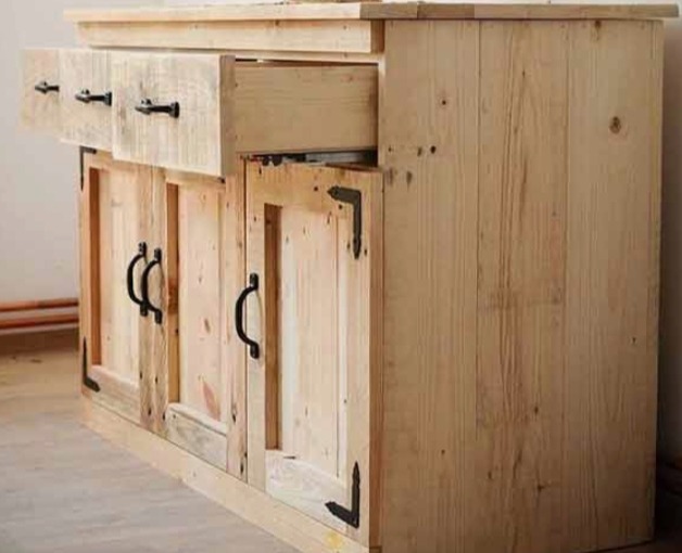 Drewniana komoda z szufladami i szafkami, wykonana z jasnego drewna z widocznymi słojami. Czarne uchwyty i metalowe wzmocnienia narożników dodają rustykalnego charakteru.