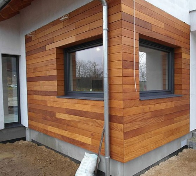 Elewacja budynku wykończona poziomymi deskami z drewna egzotycznego, z widocznymi oknami w ciemnych ramach i rurą spustową. Ujęcie z dołu, podkreślające fakturę drewna.