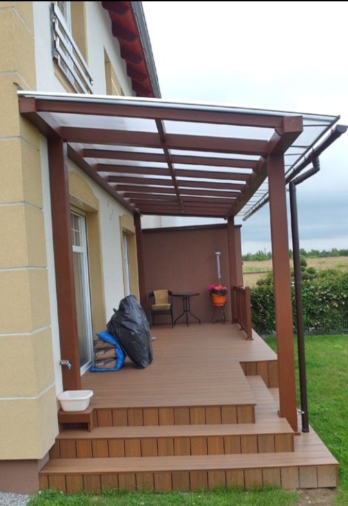 Zadaszony taras z brązową konstrukcją i drewnianymi schodami. Widoczne meble ogrodowe i rośliny w donicach. Zewnętrzna część domu z elementami dekoracyjnymi.