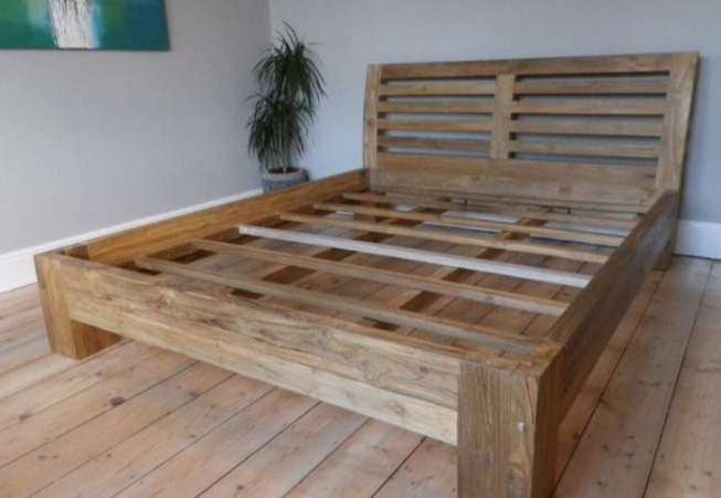 Drewniane łóżko z ażurowym zagłówkiem, wykonane z jasnego drewna o widocznej strukturze słojów. Minimalistyczny design, surowy styl, na drewnianej podłodze, z rośliną w tle.