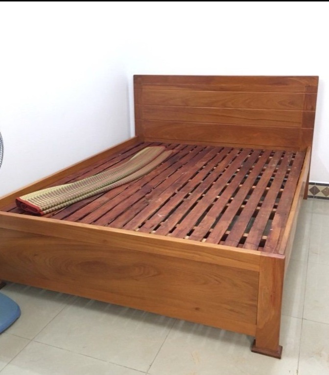 Drewniane łóżko z listwowym stelażem i wezgłowiem, na podłodze z płytek. Widoczny fragment maty na stelażu. Prosta forma, ciepły odcień drewna.