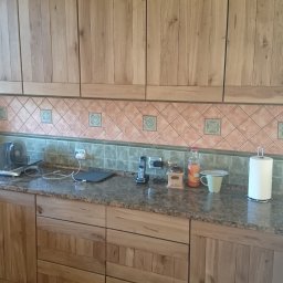 Stolmeb sp.zo.o. - Jasne, drewniane szafki kuchenne z blatem z kamienia. Widoczne detale wykończenia i wzór płytek nad blatem. Ujęcie z perspektywy osoby stojącej w kuchni.