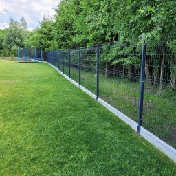 Gałązka Piotr Uslugi Sprzatajaco Ogrodnicze - Ciemnoszary płot panelowy z betonową podmurówką, otaczający zadbany, zielony trawnik z trampoliną w tle, pod pochmurnym niebem.