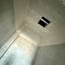 Mikołaj Ratusznik - Wnętrze łazienki w trakcie remontu: widoczne płytki na ścianach i podłodze, sufit z otworem i metalową konstrukcją, ściany przygotowane do malowania.