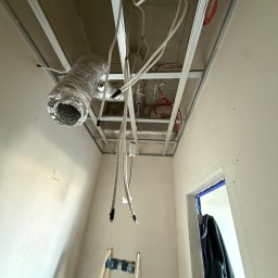 Mikołaj Ratusznik - Montaż sufitu podwieszanego z widoczną instalacją wentylacyjną i elektryczną. Metalowa konstrukcja, przewody i rura wentylacyjna w surowym wnętrzu.