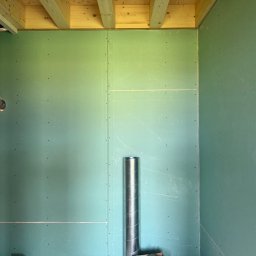Mikołaj Ratusznik - Wnętrze w trakcie wykańczania: zielone płyty gipsowo-kartonowe na ścianach, widoczne drewniane belki stropowe i fragment instalacji wentylacyjnej. Etap przed malowaniem.