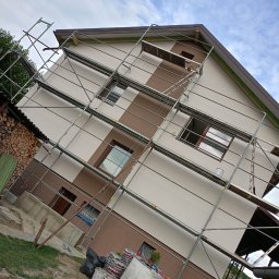 Mikołaj Ratusznik - Dwukolorowa elewacja domu w trakcie malowania, otoczona rusztowaniem. Widoczne puszki z farbą i drewno opałowe. Błękitne niebo w tle.