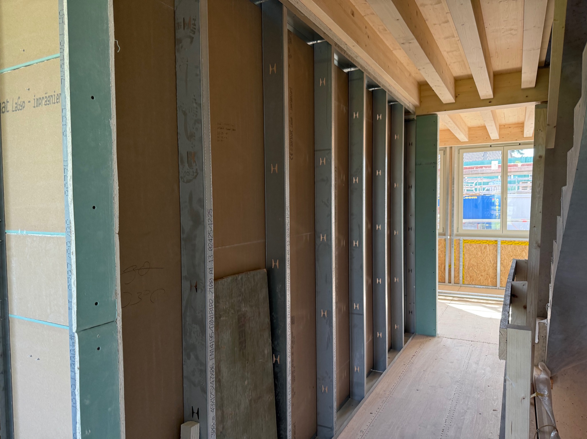 Wnętrze w trakcie budowy: metalowe profile ścian działowych z płytami gipsowo-kartonowymi, drewniany strop, okno w tle. Widok na surowy stan wykończenia.