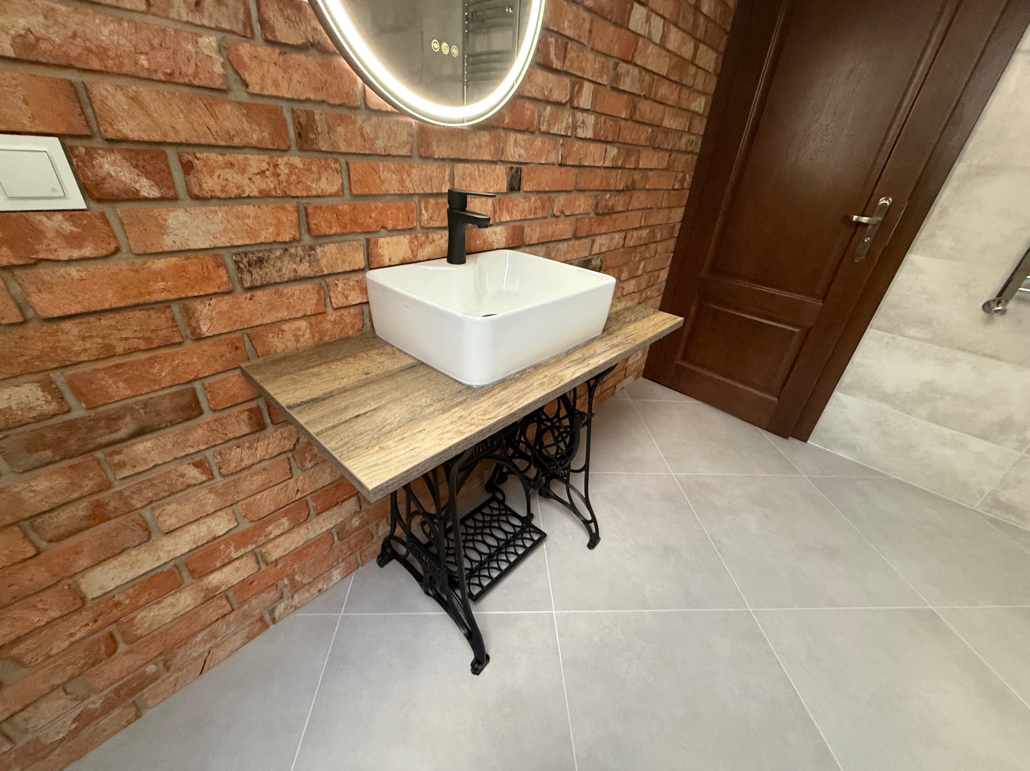 Nowoczesna łazienka z umywalką nablatową na postumencie z maszyny do szycia, ceglana ściana i okrągłe lustro z podświetleniem LED.