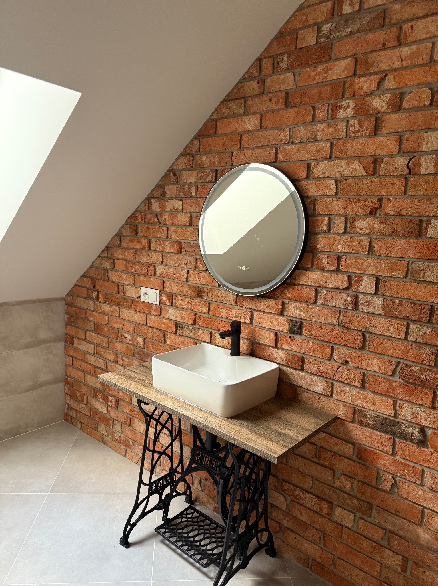 Loftowa łazienka na poddaszu: ceglana ściana, okrągłe lustro z podświetleniem i umywalka na postumencie z maszyny do szycia. Nowoczesny design z industrialnym akcentem.