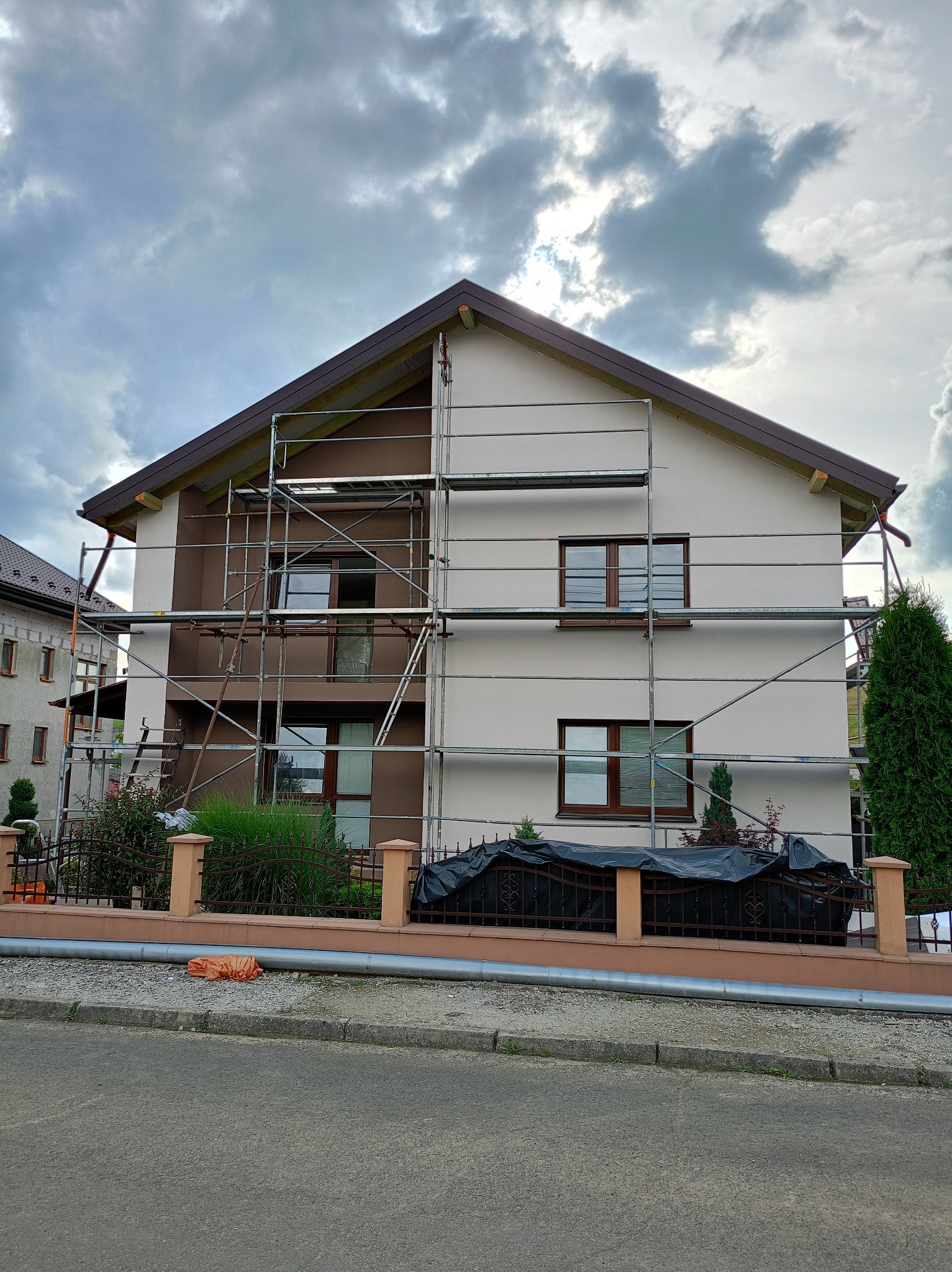 Dwukondygnacyjny dom w trakcie malowania elewacji, z widocznymi rusztowaniami i częściowo pomalowaną fasadą w odcieniach beżu i brązu, pod pochmurnym niebem.