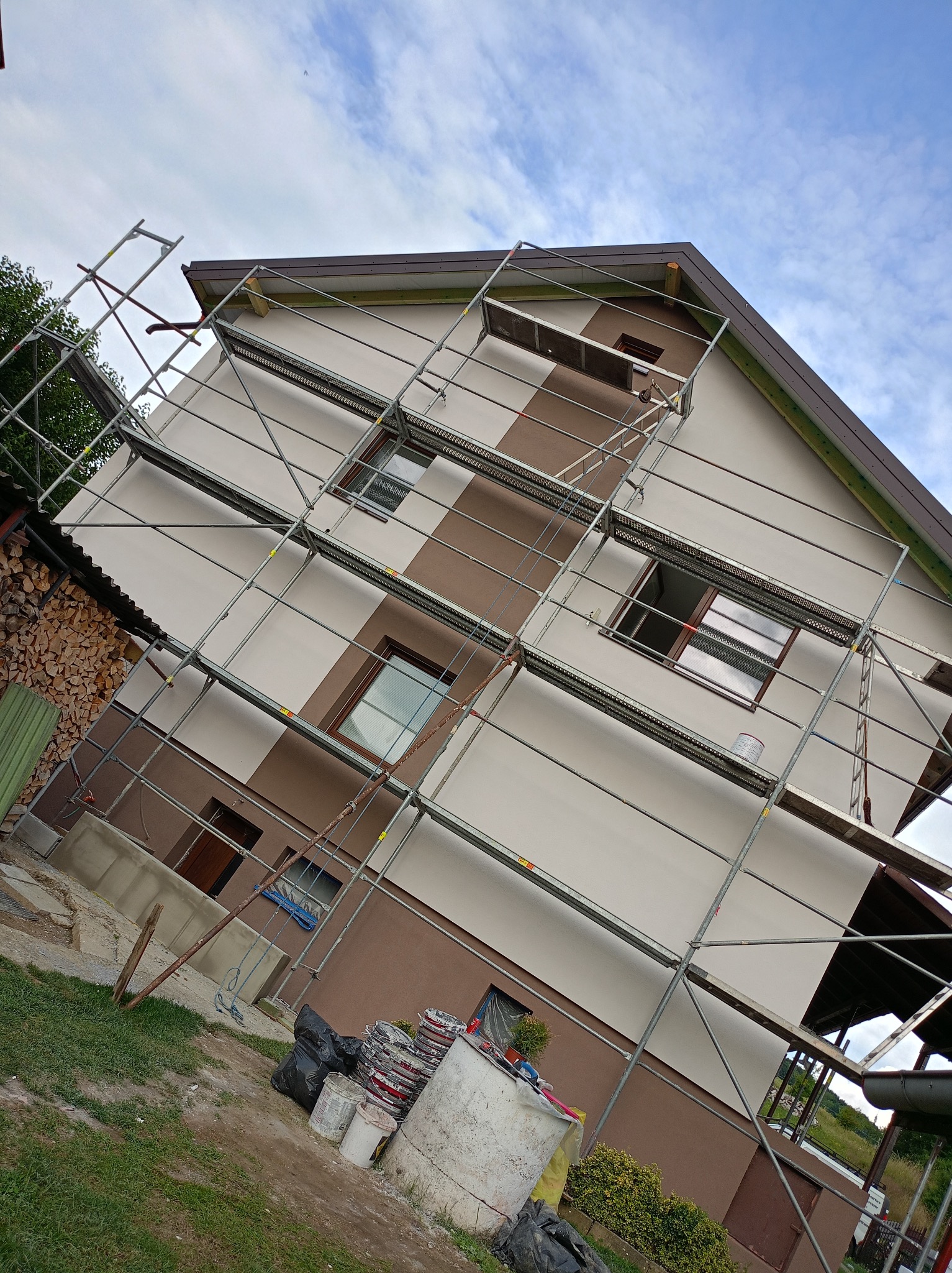 Dwukolorowa elewacja domu w trakcie malowania, otoczona rusztowaniem. Widoczne puszki z farbą i drewno opałowe. Błękitne niebo w tle.