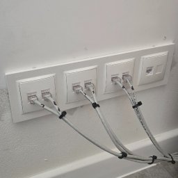 Robert Breza - Cztery białe gniazda sieciowe zamontowane w ścianie, z podłączonymi kablami Ethernet spiętymi czarnymi opaskami zaciskowymi. Widoczne fragmenty podłogi i listwy przypodłogowej.