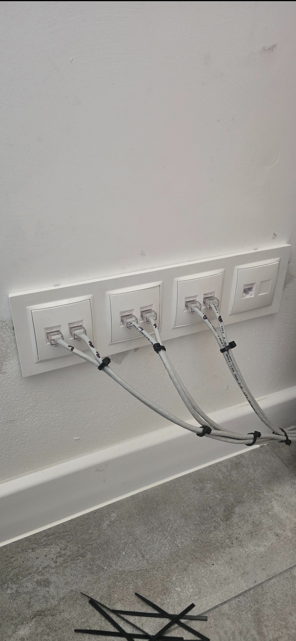 Cztery białe gniazda sieciowe zamontowane w ścianie, z podłączonymi kablami Ethernet spiętymi czarnymi opaskami zaciskowymi. Widoczne fragmenty podłogi i listwy przypodłogowej.