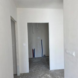 Bartosz Nowicki - Wnętrze w trakcie prac wykończeniowych: gładkie, białe ściany, widoczne instalacje wodne i elektryczne, surowa podłoga. Widok z perspektywy wejścia do pomieszczenia.
