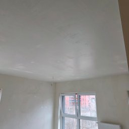 Bartosz Nowicki - Wnętrze pokoju w trakcie prac wykończeniowych. Widoczne ściany i sufit po szpachlowaniu, przygotowane do dalszych prac. Okno z widokiem na zewnątrz, grzejnik.