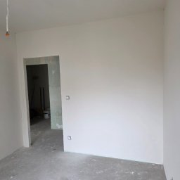 Bartosz Nowicki - Wnętrze pokoju po świeżym malowaniu na biało, widoczne gładkie ściany, surowa podłoga i otwór drzwiowy. W tle wiadro z farbą i narzędzia malarskie.