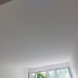 Bartosz Nowicki - Jasne wnętrze po remoncie: świeżo pomalowany sufit i ściany w bieli, widok na zieleń za oknem. Minimalistyczny design, otwarte okno.