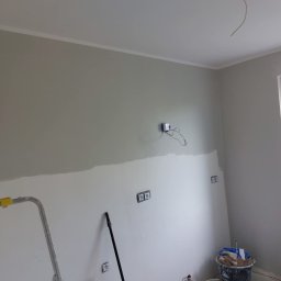 Bartosz Nowicki - Malowanie ścian w trakcie: szara farba na górze, biała szpachla na dole. Widoczne narzędzia, drabina i puszki elektryczne. Etap prac wykończeniowych.