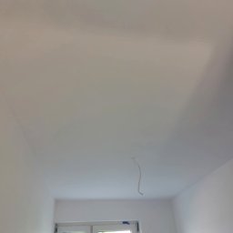 Bartosz Nowicki - Białe, świeżo pomalowane wnętrze z oknem i wystającym kablem z sufitu. Widoczne ślady taśmy malarskiej na oknie. Minimalistyczny kadr, jasne światło.