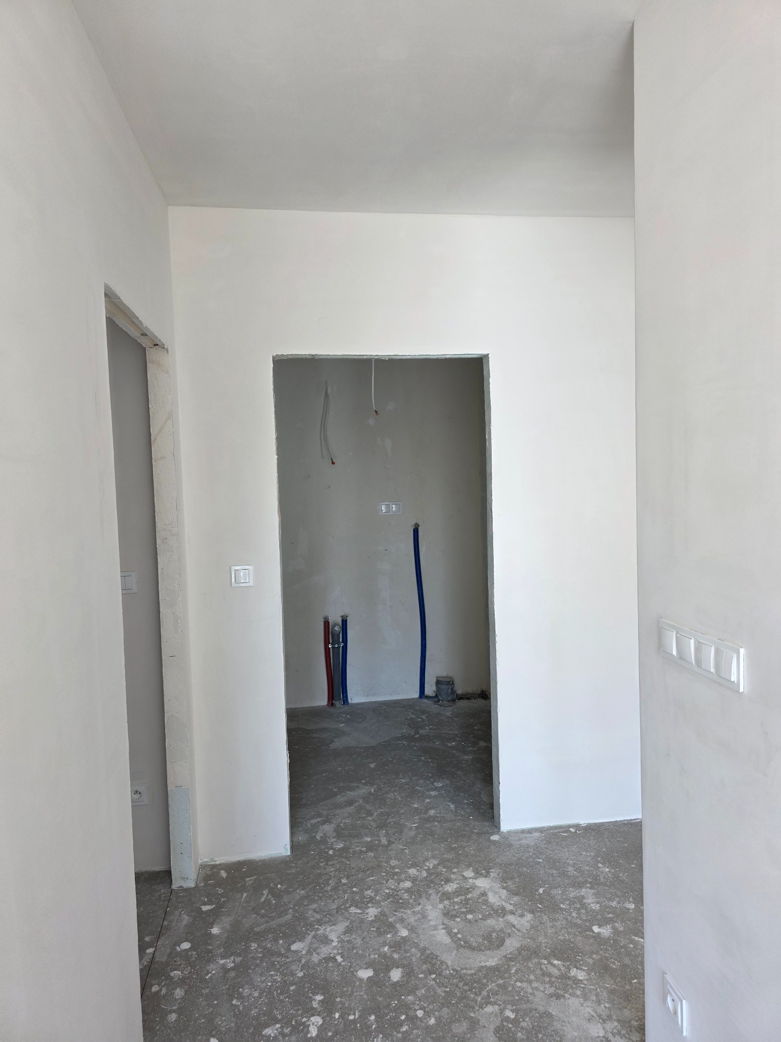 Wnętrze w trakcie prac wykończeniowych: gładkie, białe ściany, widoczne instalacje wodne i elektryczne, surowa podłoga. Widok z perspektywy wejścia do pomieszczenia.