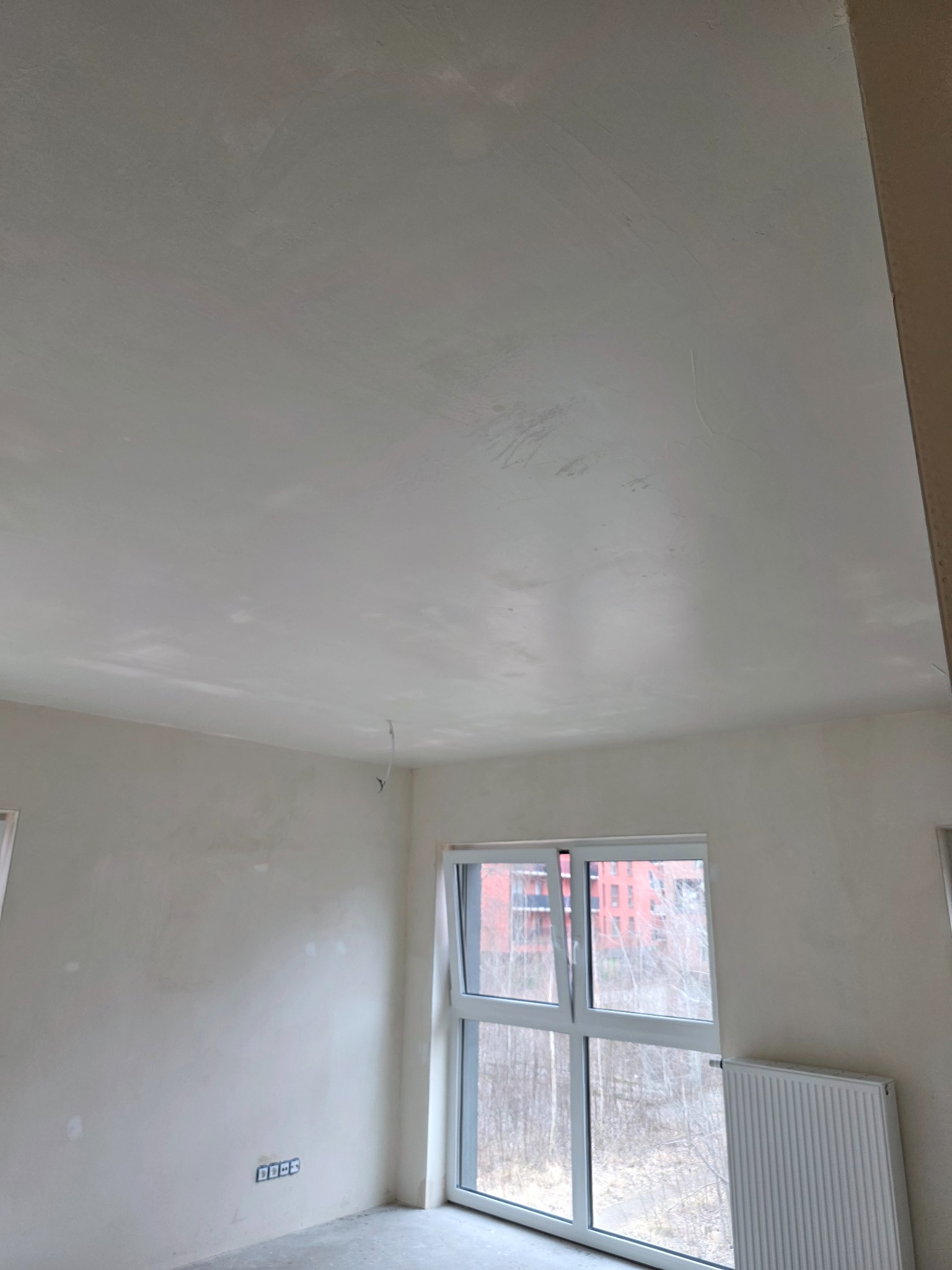 Wnętrze pokoju w trakcie prac wykończeniowych. Widoczne ściany i sufit po szpachlowaniu, przygotowane do dalszych prac. Okno z widokiem na zewnątrz, grzejnik.