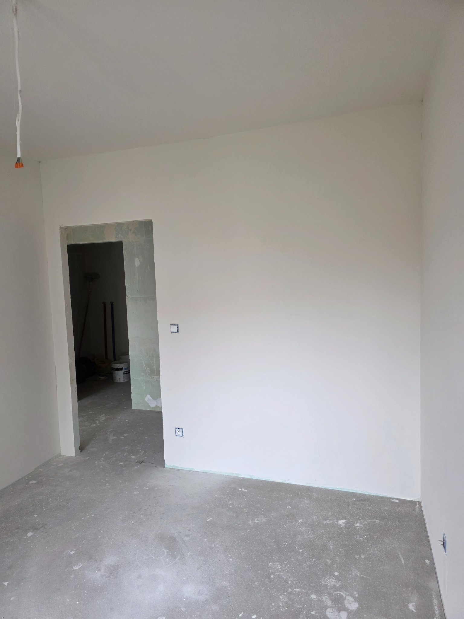 Wnętrze pokoju po świeżym malowaniu na biało, widoczne gładkie ściany, surowa podłoga i otwór drzwiowy. W tle wiadro z farbą i narzędzia malarskie.