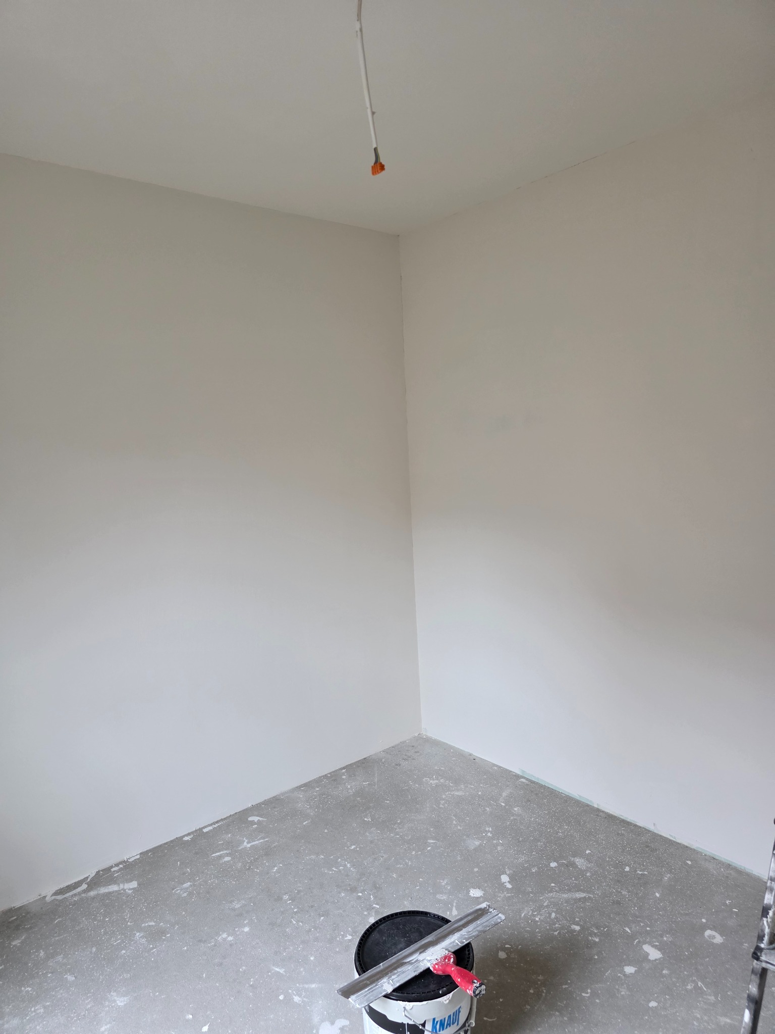 Wnętrze pokoju po nałożeniu gładzi, widoczny kubeł z masą szpachlową i szpachelka. Ściany gładkie, przygotowane do malowania. Kabel zwisa z sufitu.