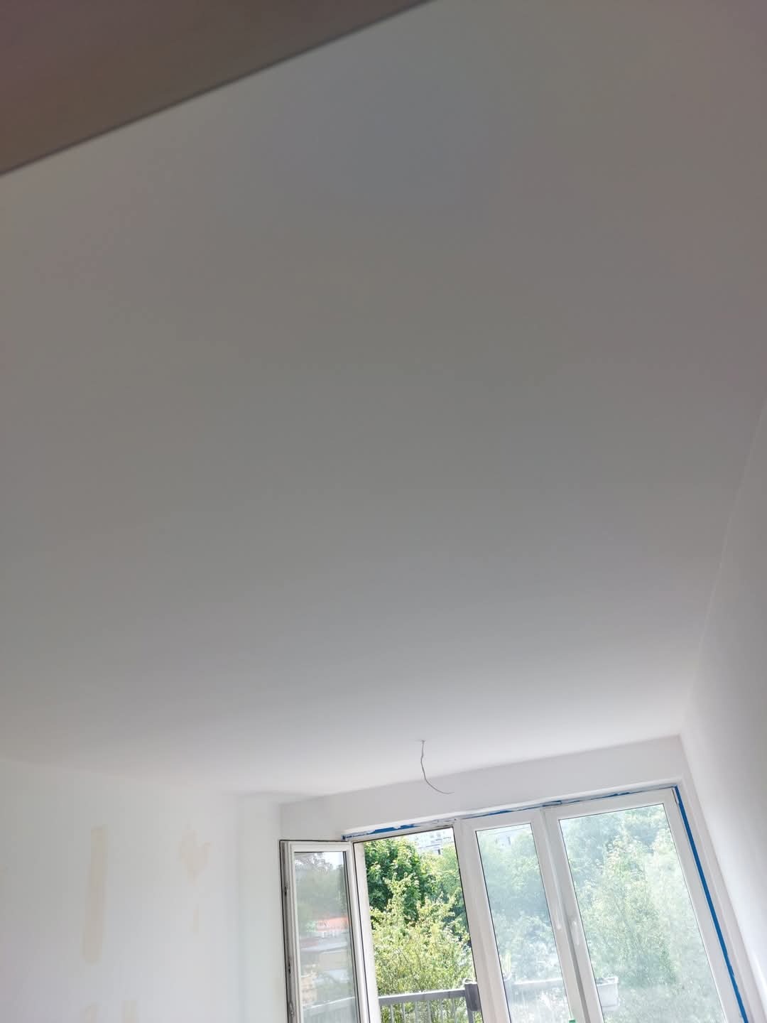 Jasne wnętrze po remoncie: świeżo pomalowany sufit i ściany w bieli, widok na zieleń za oknem. Minimalistyczny design, otwarte okno.