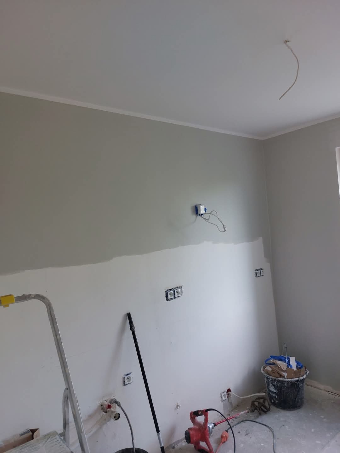 Malowanie ścian w trakcie: szara farba na górze, biała szpachla na dole. Widoczne narzędzia, drabina i puszki elektryczne. Etap prac wykończeniowych.