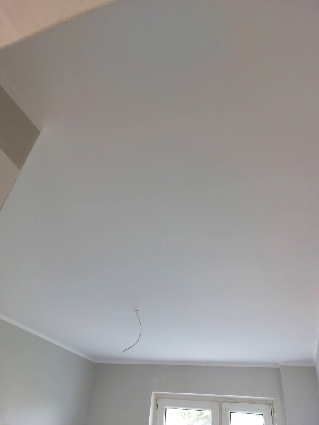 Świeżo pomalowane na biało ściany i sufit w pokoju z oknem, widoczny fragment listwy przypodłogowej i wystający kabel ze ściany. Minimalistyczne wykończenie wnętrza.