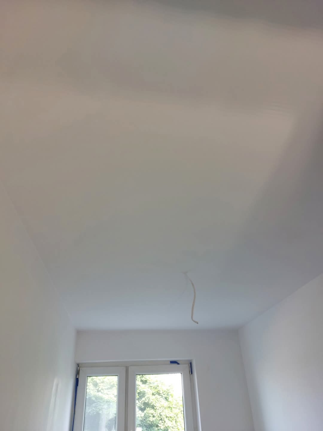 Białe, świeżo pomalowane wnętrze z oknem i wystającym kablem z sufitu. Widoczne ślady taśmy malarskiej na oknie. Minimalistyczny kadr, jasne światło.