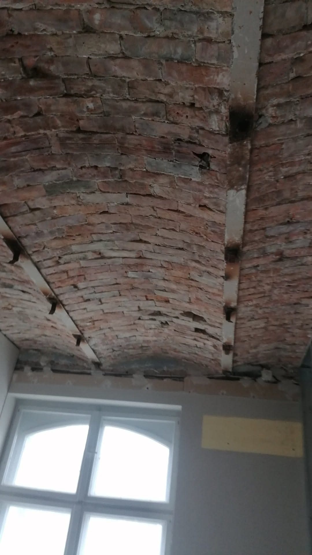 Ceglane sklepienie łukowe z metalowymi wzmocnieniami w trakcie prac remontowych, widoczne okno i fragment ściany. Charakterystyczna struktura cegieł i oświetlenie.
