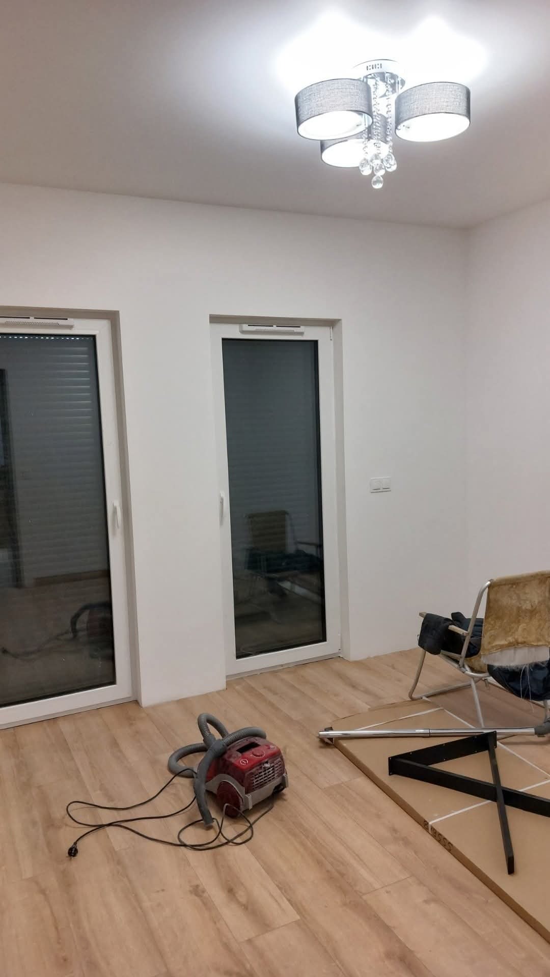 Jasne wnętrze z panelami podłogowymi, odkurzaczem i elementami mebli do montażu. Ściany świeżo pomalowane na biało, widoczne dwa okna balkonowe.