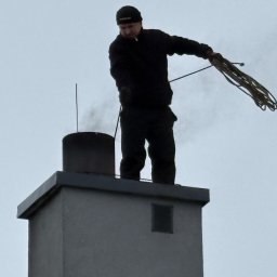 R.K.Kominiarstwo - Kominiarz na dachu komina z dymem, trzymający linę. Ujęcie z dołu, pochmurne niebo w tle. Widoczny fragment czapki kominiarza.