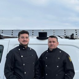 R.K.Kominiarstwo - Dwóch kominiarzy w czarnych uniformach stoi przed białym busem z drabiną i cylindrem na dachu, prezentując tradycyjny strój i gotowość do pracy.