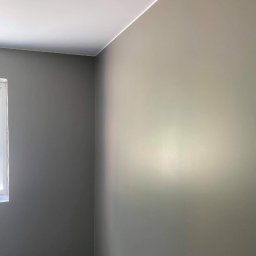 Mateusz Górski - Pomieszczenie po malowaniu ścian na szaro, widoczne okno i fragment podłogi zabezpieczonej folią malarską. Ściany gładkie, sufit biały. Ujęcie z rogu pokoju.